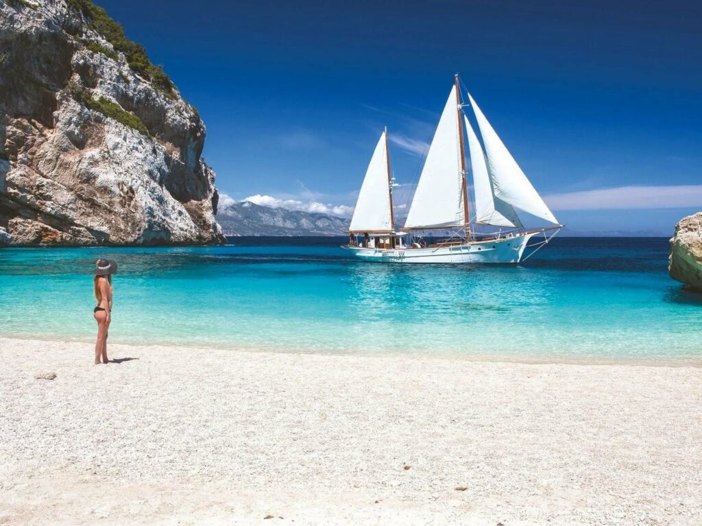 Donna in costume in spiaggia che guarda una barca a vela bianca navigare nel mare turchese della Sardegna