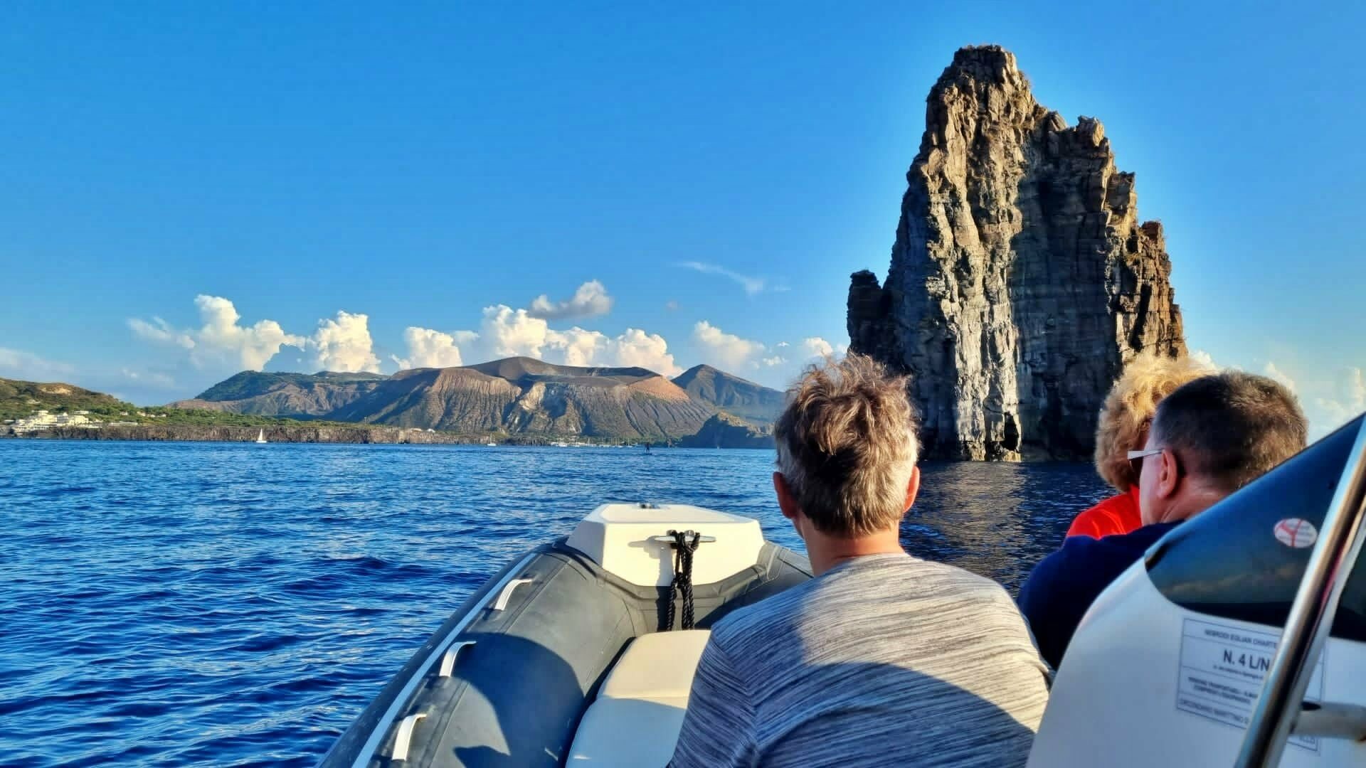 Persone a bordo di una barca partecipano a un escursione alle Isole Eolie e davanti a loro vedono un faraglione nel mare