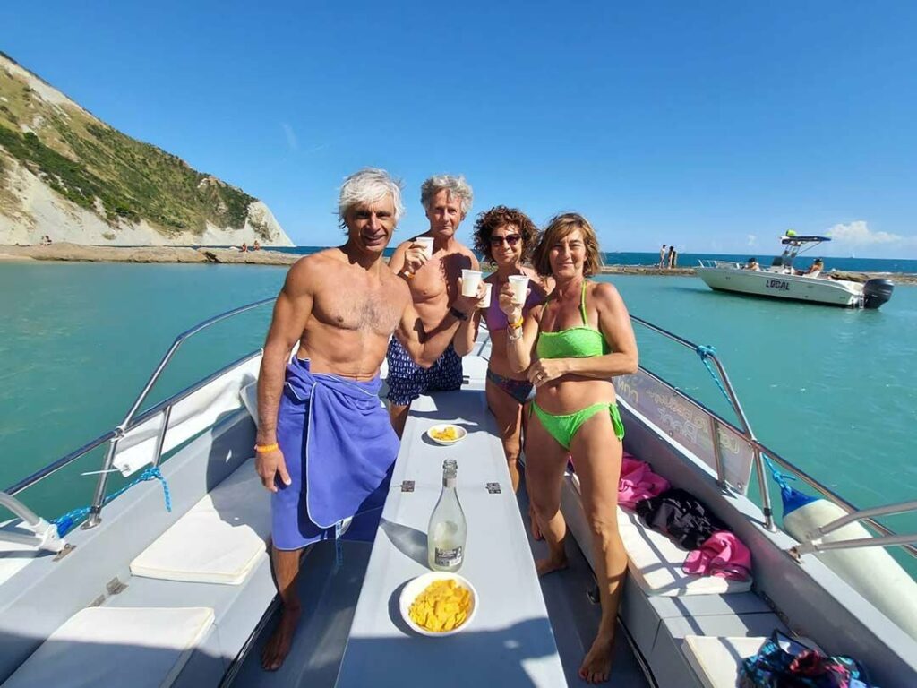 Gruppo di uomini e donne brinda in barca con un aperitivo con il mare e le scogliere della Riviera del Conero sullo sfondo