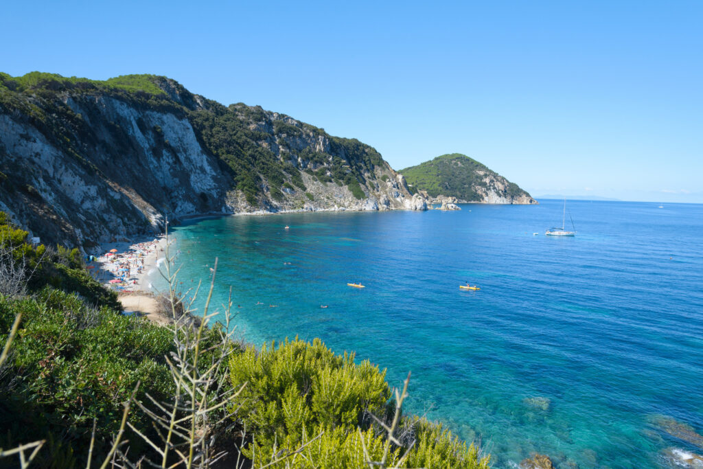 Panorama della spiaggia di sabbia bianca sull'Isola d'Elba con acqua cristallina, barche a vela e kayak al largo, circondata da scogliere verdi e mare turchese