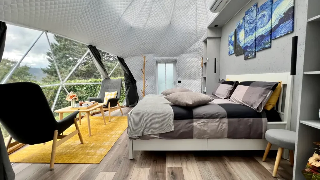Interno di bubble room con vetrate panoramiche: letto matrimoniale, area relax e vista su verde e colline