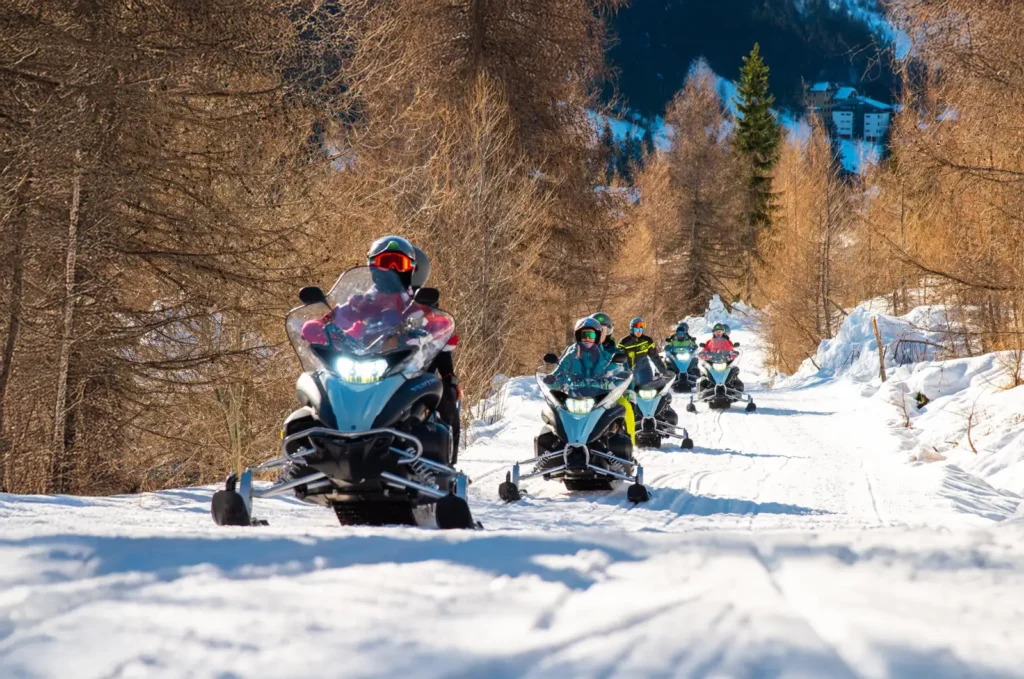 scursione in motoslitta a Madesimo in inverno, gruppo in fila su sentiero innevato tra larici e paesaggio alpino