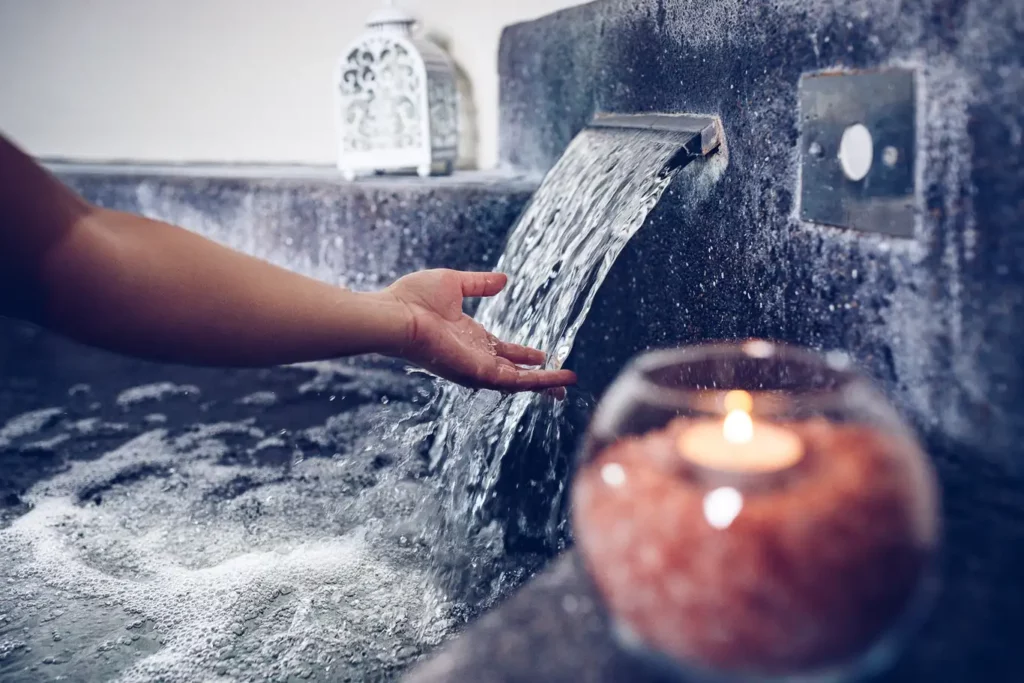 Mano sotto getto d’acqua a cascata in vasca idromassaggio con candela accesa, atmosfera sensoriale per una giornata in spa nelle Marche
