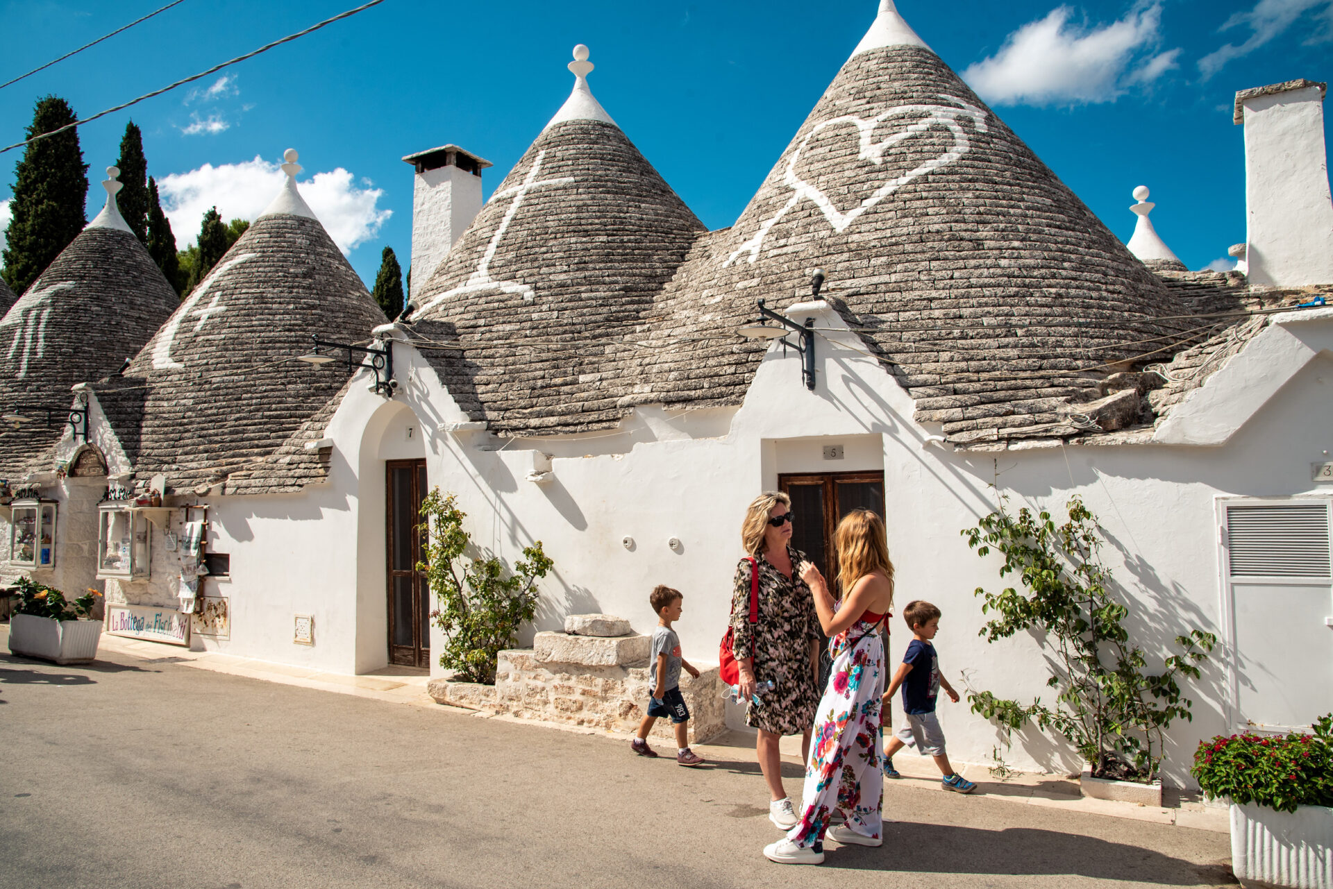 Trulli di Alberobello in Puglia: case bianche con tetti conici, due donne e bambini che passeggiano nel borgo sotto cielo azzurro.