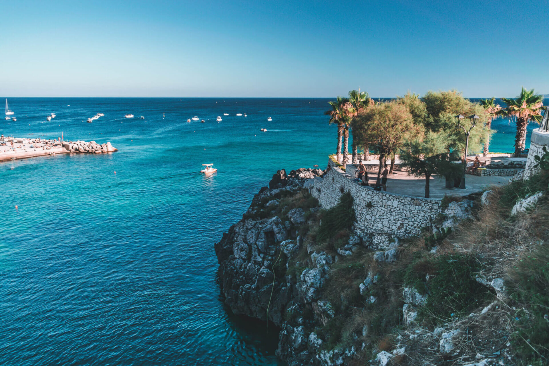 Promontorio roccioso con palme affacciato sul mare turchese del Salento con barche ormeggiate nella baia