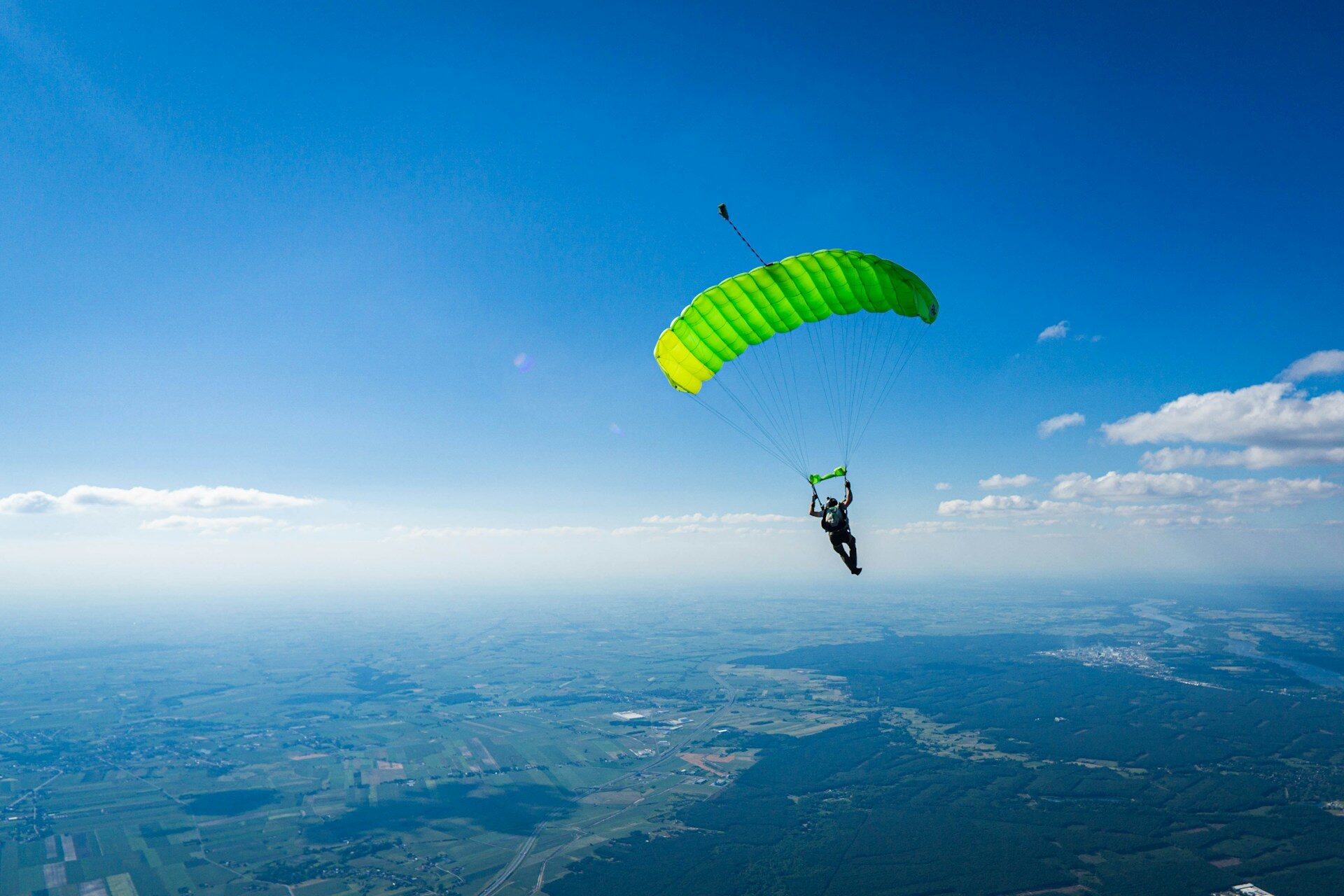 Vista aerea di un paracadutista in volo con un paracadute verde brillante contro un cielo blu limpido e un vasto panorama di campi sullo sfondo.