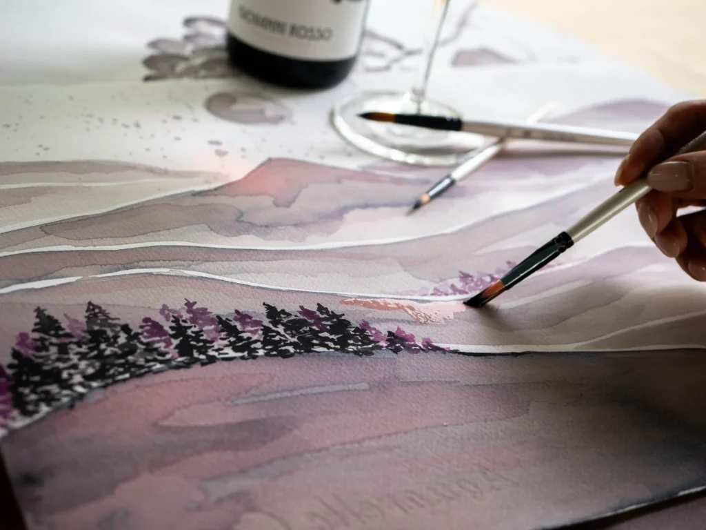 Laboratorio di wine painting a Serralunga d'Alba in Piemonte, con una mano che dipinge un paesaggio usando il vino Barbera accanto a un calice.