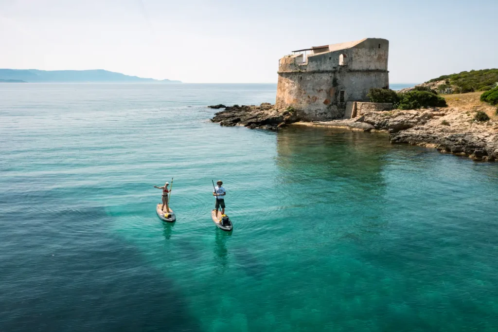 Due persone in SUP sulle acque turchesi della costa di Alghero con la Torre del Lazzaretto sullo sfondo