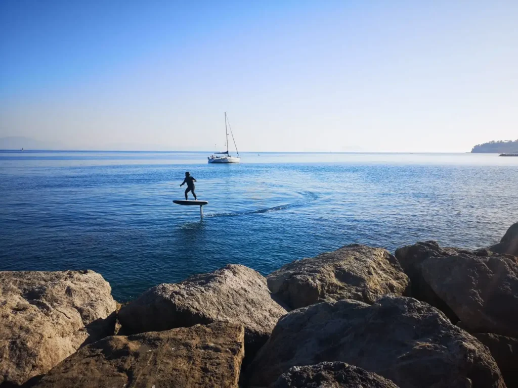 Persona in piedi su un eFoil nel Golfo di Napoli con barca a vela sullo sfondo