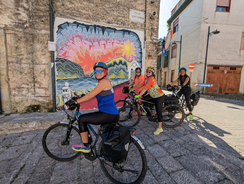 Due cicliste in e-bike davanti a un murales a Piana degli Albanesi